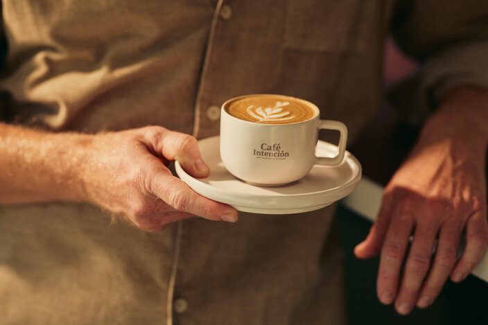 Een barista serveert een kopje koffie met latte art bij Café Intención, waarbij de rijke crema mooi tot zijn recht komt in een stijlvol kopje met schoteltje.