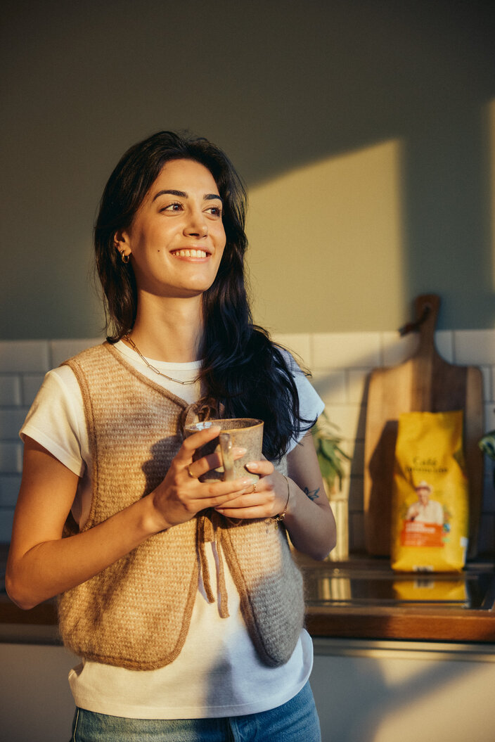 A woman smiles warmly while holding a ceramic cup in a bright kitchen, with coffee bags visible in the background.
