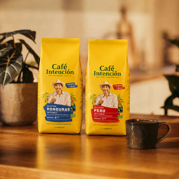 Torby z kawą Café Intención z Hondurasu i Peru na drewnianym stole, a w tle filiżanka kawy i rośliny.